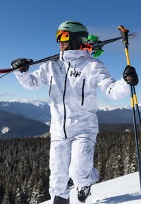 Combinaison de ski camouflage blanc avec des accents noirs, associée à un casque vert et des lunettes de ski miroir, portant des skis, sur un fond de montagne enneigée.