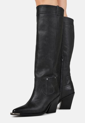 Bottes de cowboy hautes en cuir noir avec des bouts pointus, des embouts en métal, des fermetures éclair latérales et des talons blocs portées par une personne.
