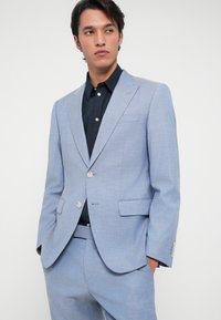 Šviesiai mėlynas švarko striukė iš tekstūruotos medžiagos su įpjovimu, dviem priekinėm kišenėm ir vieno mygtuko užsegimu. Derinama su juodu marškiniu.