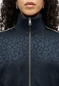 Μπλε λεοπάρ μπουφάν με ψηλό γιακά, κλείσιμο με φερμουάρ και λευκές πλαϊνές ρίγες. Υφασμάτινη υφή και μεταλλική λεπτομέρεια φερμουάρ.