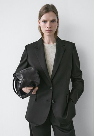 Kvinde med glat hår iført sort blazer og bukser, holder en sort lædertaske foran en ensfarvet hvid væg.