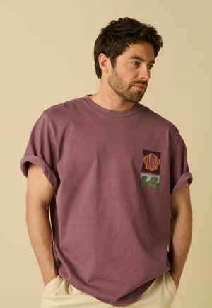 Man draagt een mauve T-shirt met opgerolde mouwen en een patch van een schelp en golf op de borst, beige broek, kijkt opzij tegen een beige achtergrond.