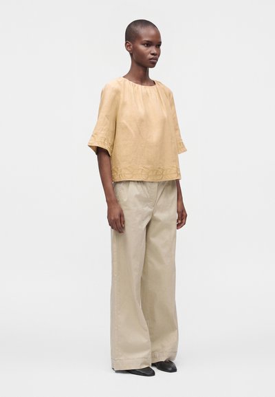Femme debout portant un haut beige brodé ample avec de larges manches courtes et un pantalon large beige clair avec des chaussures noires.