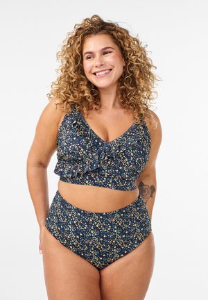 Femme souriante aux cheveux bouclés portant un maillot deux pièces bleu à fleurs avec des volants et un tatouage de rose sur son bras gauche.