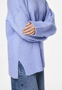 Lichten lavendelkleurige gebreide trui met geribde manchetten, zijsplitten en een relaxte pasvorm, gecombineerd met donkerdenim jeans. Zachte textuur zichtbaar.