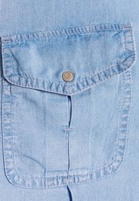 Ljusblå denimskjorta med en ficka med klaff och silverfärgad tryckknapp, förstärkt sömnad och en slät textur.