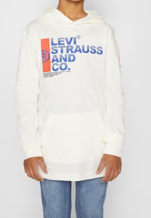 Młoda osoba ubrana w białą bluzę z kapturem Levi Strauss z niebiesko-czerwonym logo, w połączeniu z niebieskimi dżinsami, stojąca na tle jednolitego tła.