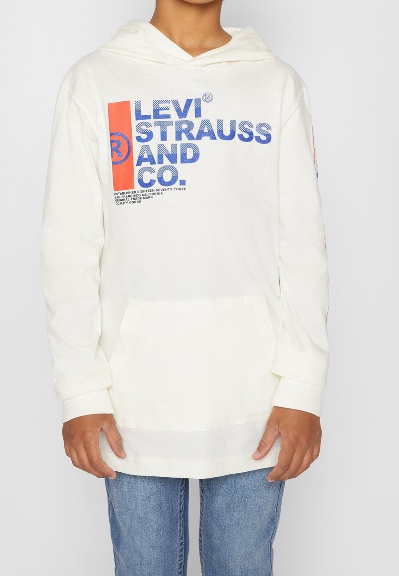 Młoda osoba ubrana w białą bluzę z kapturem Levi Strauss z niebiesko-czerwonym logo, w połączeniu z niebieskimi dżinsami, stojąca na tle jednolitego tła.