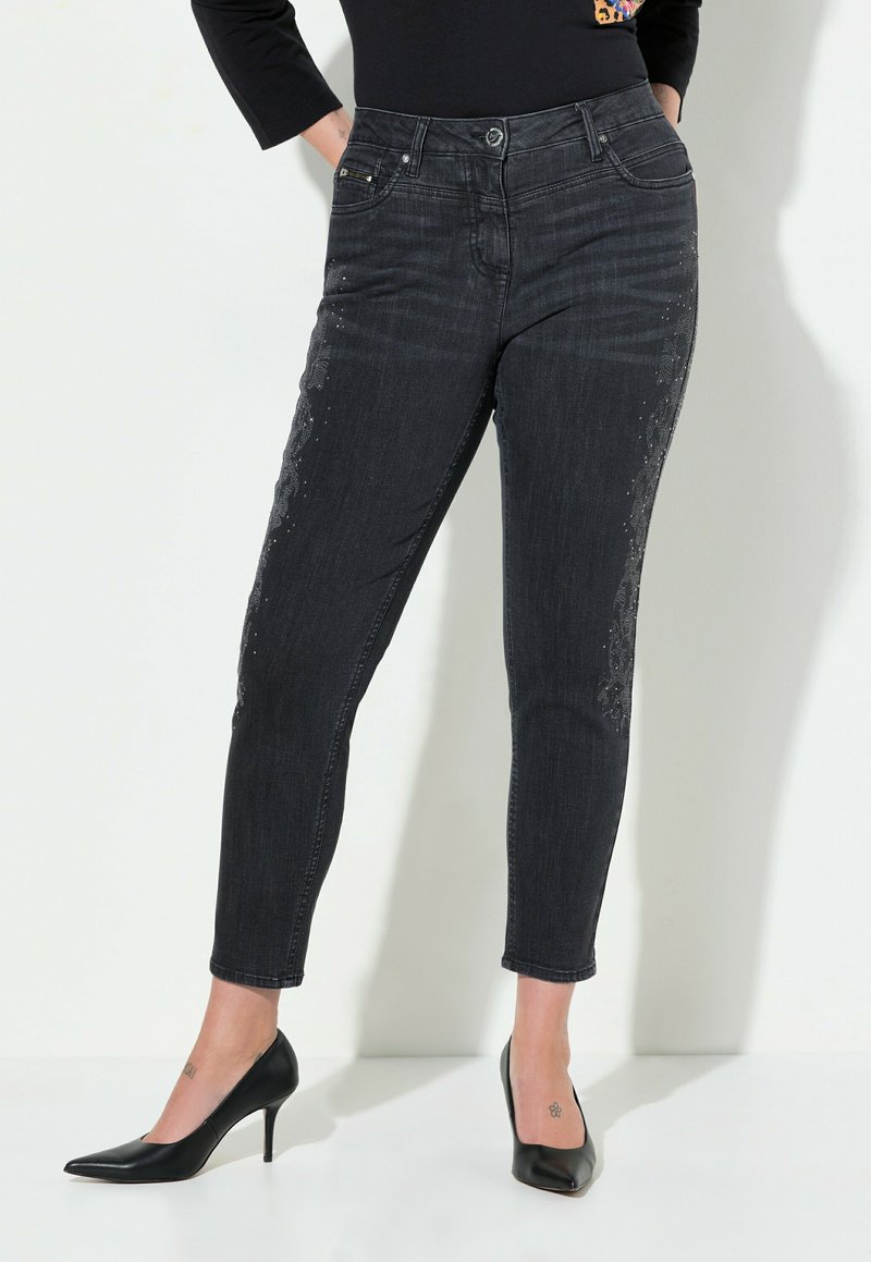 Jeans de mezclilla negra con un diseño ajustado, que presentan rhinestones decorativos en el costado y una cintura de media altura. Combinados con tacones altos negros.