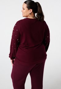 Vrouw met paardenstaart, in een bordeauxrode trui met parelaccenten op de mouwen en bijpassende textuurbroek, zijwaarts gekeerd tegen een witte achtergrond.