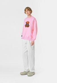 Sudadera rosa con un gráfico de oso de peluche, combinada con pantalones blancos que tienen cordones y un detalle de cadena, y calzado gris.