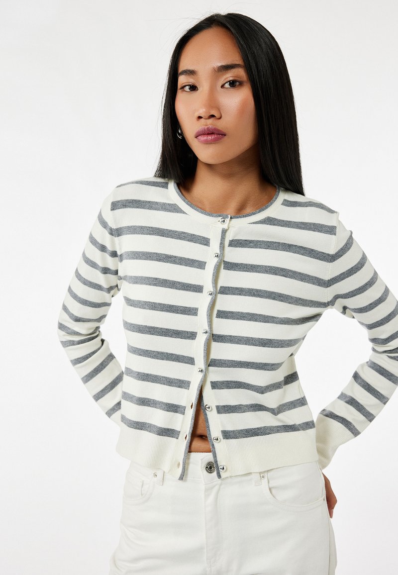 Grijze en witte gestreepte cardigan van zachte breiwerk, met een ronde hals en een knoopsluiting, gecombineerd met witte broeken.