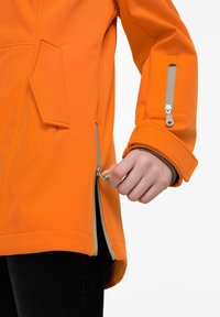 Oranssi vedenpitävä takki, jossa on vetoketjusuljenta, sivutaskut ja joustavat hihansuut. Kangas on sileä, ja siinä on näkyviä sauman yksityiskohtia.