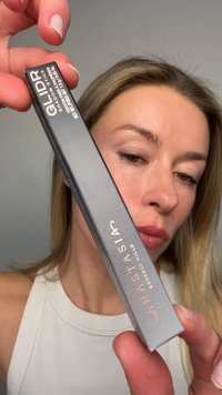 Femme tenant une boîte grise étiquetée Anastasia Beverly Hills GLIDR shadow stick près de son visage sur un fond uni.