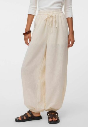 Femme portant un pantalon ample beige clair avec cordon de serrage et poignets élastiqués, assorti à des sandales marron foncé et un haut blanc à manches longues.