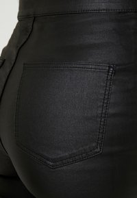 Svarta, högmidjade byxor i slät tyg. Har en enda bakficka med sydd kant, ingen synlig logotyp.