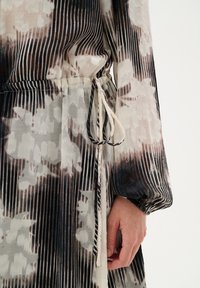 Vestido maxi de tejido ligero a rayas con un patrón floral, en tonos neutros de beige y negro, con cintura ajustada y detalle de lazada.