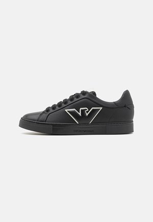 Zapatilla negra de cuero con un acabado suave, que presenta un prominente logo plateado en el lateral, puntera redonda y suela de goma texturizada.
