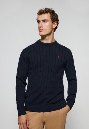 Donkerblauwe kabelgebreide trui met ronde hals, voorzien van geribbelde manchetten en zoom. Een klein rood logo-accent is zichtbaar op de borst.