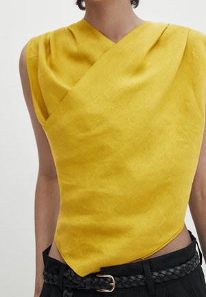 Ärmelloses gelbes Leinen-Top mit asymmetrischem Ausschnitt und spitzem Saum, getragen mit schwarzem geflochtenem Gürtel und schwarzer Hose.