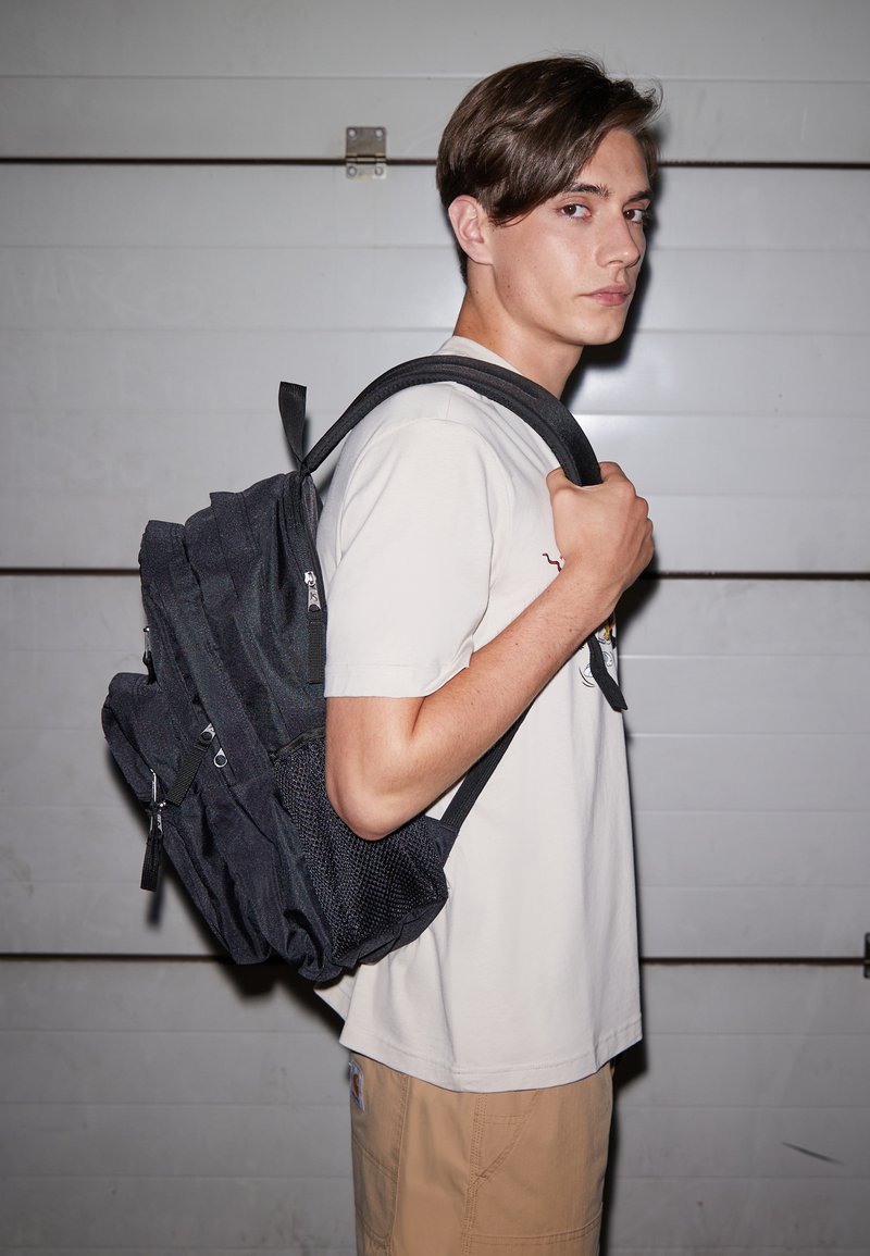 Mochila negra texturizada con múltiples bolsillos con cremallera y un bolsillo lateral de malla, llevada sobre una camisa clara y pantalones cortos khaki.