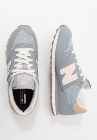 Zapatillas deportivas grises con material de malla y sintético, con un logo "N" rosa, detalles en crema y suela de goma marrón.