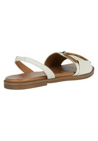 Vit platt sandal med brun sula, ankelrem och guldbuckledetalj på den främre remmen, sidovy.