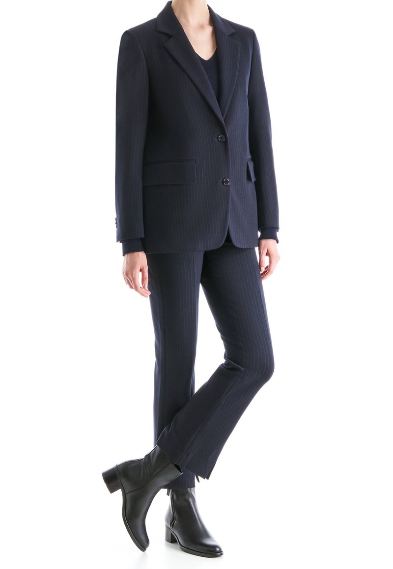 Personne portant un costume rayé sombre avec une veste à deux boutons, un pantalon assorti, des bottes noires, debout avec une jambe croisée sur l'autre.