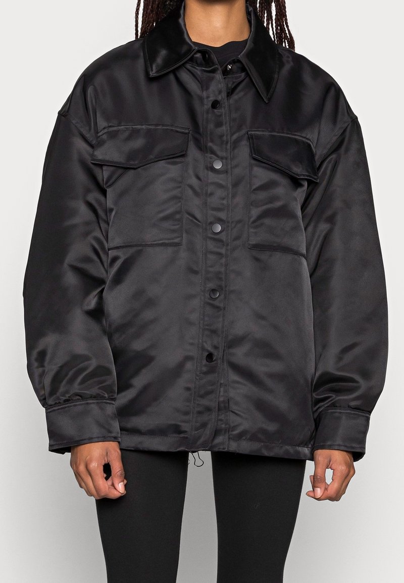 Black satin oversized jacket with a pointed collar, two chest pockets, front buttons, and elasticized cuffs. Smooth texture and relaxed fit.