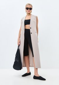 Dlouhá béžová bezrukávová vesta, černý crop top, černé legíny ke kolenům, ploché černé boty, černá tkaná taška, doplněno zlatou choker náhrdelníkem a slunečními brýlemi.