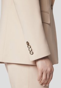 Jasno beżowa marynarka o dopasowanym kroju z teksturowanym wykończeniem. Cztery guziki w odcieniu złota na mankietach, gładka klapa oraz usztywnione ramiona.