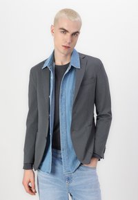 Grau Blazer über einem hellblauen Denimhemd, kombiniert mit einem schwarzen T-Shirt und hellblauen Jeans. Merkmale ein körperbetontes Design und minimale Details.