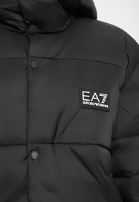 Čierna pérová bunda s kapucňou, s prešitým vzorom a nášivkou loga EA7 Emporio Armani na hrudi. Plastové cvoky.