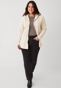 Helle beigen Zip-Hoodie mit einer weichen Textur, kombiniert mit dunklen Jeans und Ankle Boots. Fokus auf lässiger Passform und praktischen Designelementen.