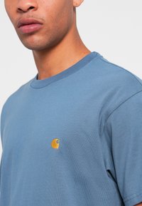 T-shirt en coton bleu clair à col rond, avec un petit logo orange sur la poitrine et des manches courtes. Texture lisse, design minimal.