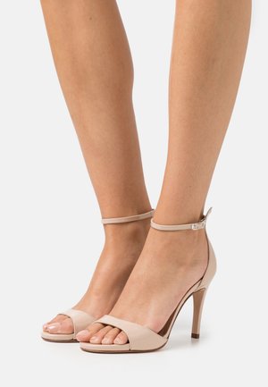 Des pieds portant des sandales à talons hauts en cuir verni nude, avec de fines brides autour de la cheville et des orteils ouverts, présentés sur fond blanc uni.