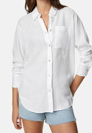 Mujer que lleva una camisa blanca suelta abotonada con un bolsillo en el pecho y shorts vaqueros azul claro con bordes deshilachados.