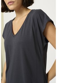 Femme portant un t-shirt gris foncé à manches courtes et col en V, vue des épaules jusqu'au milieu du torse sur un fond clair uni.