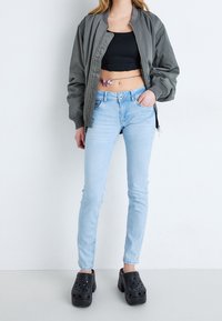 Šedá bomber bunda, černý crop top, světle modré skinny džíny a černé platformové dřeváky. Výrazný pásek s motýlem jako akcent. Přítomné hladké textury.