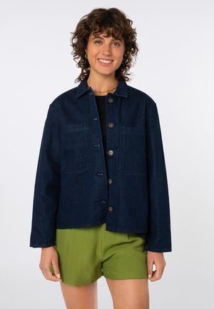 Woman with curly hair smiling, wearing a dark blue buttoned denim jacket over a black top and green shorts against a white background.