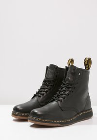 Par de botas de cuero negro con cordones y costuras amarillas, y tiradores de marca, sobre una superficie y fondo blancos.