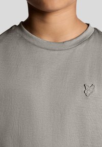 Lysegrå t-shirt lavet af blødt stof, med rund hals og et lille broderet logo på venstre bryst. Glat tekstur.