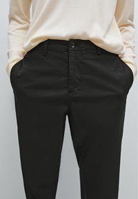 Chino neri, leggeri e dalla texture liscia, con chiusura a bottone e tasche laterali, abbinati a un top beige a maniche lunghe.