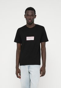 Μαύρο βαμβακερό μπλουζάκι με ορθογώνιο κόκκινο και λευκό λογότυπο στο μπροστινό μέρος. Κοντά μανίκια και κλασικός σχέδιο στρογγυλής λαιμόκοψη.