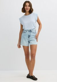 Camiseta de algodón azul claro con hombreras, combinada con pantalones cortos de denim desgastados de cintura alta. Sandalias de tiras negras completan el conjunto.