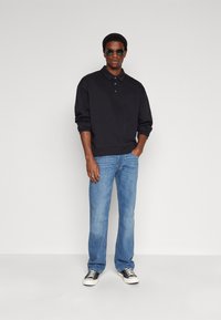 Pull noir en coton avec un col et des boutons, associé à un jean en denim bleu clair. Des baskets noir et blanc complètent la tenue.