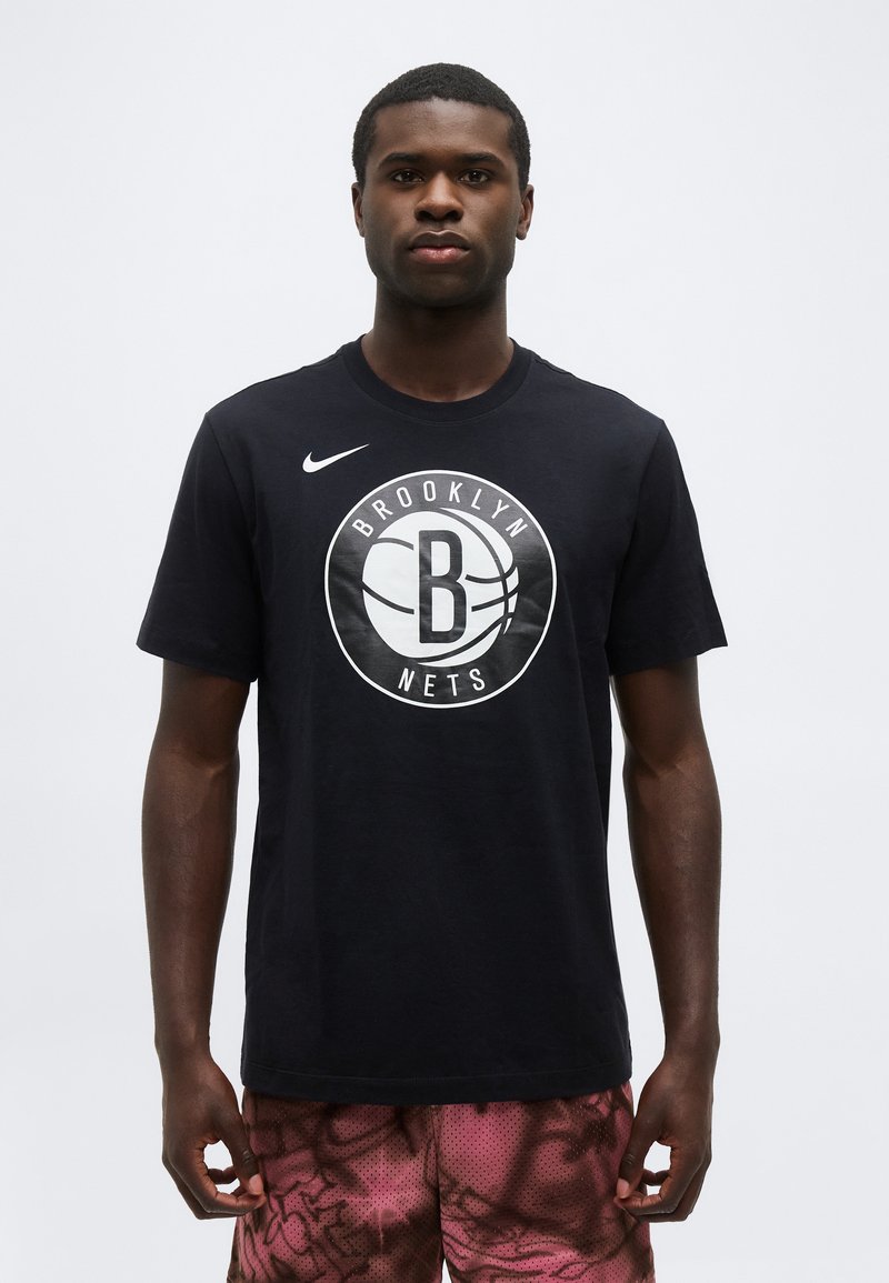 Άνδρας που στέκεται, φοράει μαύρο t-shirt Brooklyn Nets και μωβ σορτς με σχέδια, κοιτάζοντας μπροστά σε λευκό φόντο.