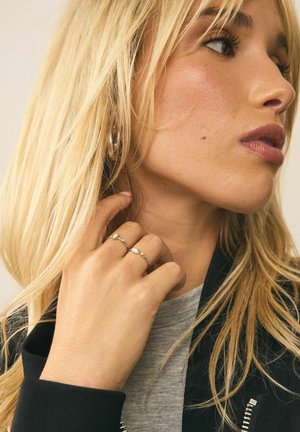 Two silver rings with a polished finish, featuring circular and teardrop shapes. The hand is partially raised, with long, wavy blonde hair.
