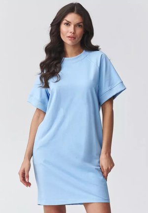 Kobieta o długich, ciemnych włosach, ubrana w jasnoniebieską, krótką, casualową sukienkę, stojąca na tle jednolitego, jasnego tła.