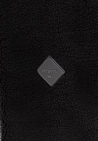 Črn flis z teksturirano površino in sivo logotipno zaklopko v obliki diamanta, ki vsebuje diagonalne črte ter ime blagovne znamke.